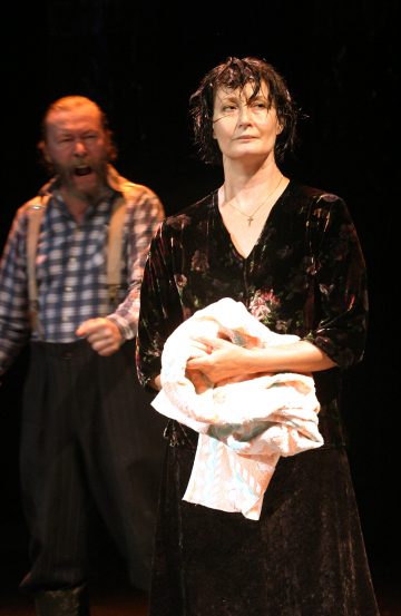 Nainen, jolla on märät hiukset ja vakava ilme, seisoo pyyhe kädessään, kun taas parrakas mies, jolla on ruudullinen paita ja henkselit, huutaa taustalla hämärästi valaistulla lavalla.