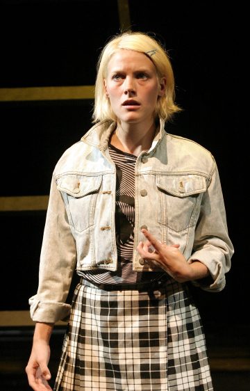 Lyhyet vaaleat hiukset omaava henkilö, jolla on päällään leikattu farkkutakki ja mustavalkoruutuinen hame, seisoo lavalla yllättynyt tai intensiivinen ilme tummaa taustaa vasten valaistuna.