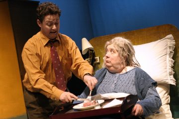 Oranssiin paitaan ja punaiseen solmioon pukeutunut henkilö yrittää leikata ruokaa tarjottimelle vanhemmalle harmaahiuksiselle naiselle, joka istuu sängyssä yllättyneen tai huolestuneen näköisenä. Kohtaus näyttää olevan näytelmästä.