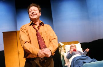 Oranssiin paitaan ja löysään solmioon pukeutunut mies hymyilee ja lyö kätensä yhteen seisoessaan lavalla; hänen takanaan sairaalasängyssä makaa vanhempi nainen, jolla on kädessään puhelimen luuri.