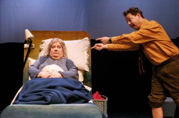Vanhempi nainen makaa sängyssä sinisillä huovilla peitettynä ja vakavana, kun oranssiin paitaan ja ruskeisiin housuihin pukeutunut mies osoittaa ja elehtii häntä vilkkaasti. Kohtaus näyttää olevan näytelmästä.