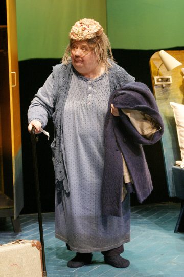 Siniseen yöpaitaan ja neuletakkiin pukeutunut iäkäs henkilö seisoo keppi kädessään, pitelee tummaa takkia toisessa käsivarressa ja kantaa kukkien koristamaa hattua. Taustalla näkyy matkalaukku ja joitakin huonekaluja.