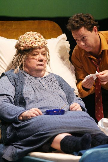 Siniseen mekkoon ja hattuun pukeutunut vanhempi nainen istuu sängyssä kulhon kanssa, kun oranssiin paitaan ja punaiseen solmioon pukeutunut mies nojaa häntä kohti nenäliina kädessään ja näyttää huolestuneelta.