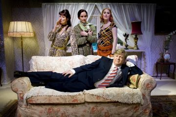 Neljä ihmistä poseeraa vintage-olohuoneessa; kolme naista seisoo taustalla, kun taas pukumies nojaa itsevarmasti etualalla olevalla kukkasohvalla. Huone on lämpimästi valaistu ja sisustettu retrotyyliin.