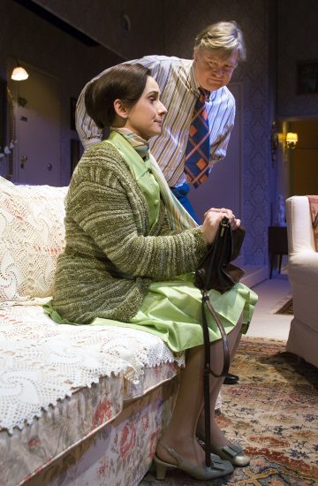 Vihreään mekkoon ja villatakkiin pukeutunut nainen istuu sohvalla käsilaukku ja keppi kädessään ja katsoo eteenpäin. Hänen takanaan seisoo raitapaitainen ja solmioinen mies, joka nojaa eteenpäin. Kohtaus näyttää sijoittuvan olohuoneeseen.