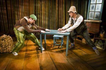 Kaksi värikkäisiin pukuihin pukeutunutta miestä nykii sinisen pöydän vastakkaisia päitä maalaismaisessa, lämpimästi valaistussa huoneessa, jossa on puulattiat ja -seinät. Toisella miehellä on vihreät housut ja lippalakki, toisella luppahattu ja liivi.