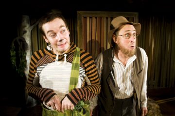 Lavalla esiintyy kaksi teatteripukuista miestä; toisella on raidallinen paita, vihreät housunkannattimet ja leikkisät kasvot, kun taas toisella on silmälasit, hattu ja liivit ja hän näyttää yllättyneeltä. Taustalla on puupaneelit.