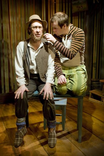 Kaksi teatteriasuihin pukeutunutta miestä istuu lähekkäin tuoleilla puisella näyttämöllä. Toisella on silmälasit, hattu ja ruudulliset tossut, ja hän katsoo eteenpäin, kun taas toinen, raidallisiin vaatteisiin pukeutunut, kumartuu sisäänpäin ja tekee eleitä korvansa lähellä.