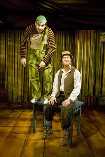 Kaksi leikkisästi pukeutunutta miestä poseeraa sisätiloissa; toinen seisoo jakkaralla vihreissä haalareissa ja raidallisessa paidassa, kun taas toinen istuu sinisellä tuolilla liivi, hattu ja silmälasit yllään. Tausta on puinen ja lämpimästi valaistu.
