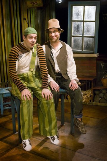 Kaksi värikkäisiin asuihin pukeutunutta miestä istuu jakkaroilla sisätiloissa. Toisella on vihreä hattu, raidallinen paita ja vihreä haalari; toisella on silmälasit, ruskehtava hattu ja liivi. He hymyilevät ikkunan vieressä viihtyisässä, maalaismaisessa ympäristössä.