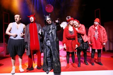 Kuusi värikkäisiin teatteripukuihin pukeutunutta näyttelijää seisoo kirkkaasti valaistulla näyttämöllä; useimmat ovat pukeutuneet punaiseen, kun taas keskimmäinen hahmo on pukeutunut mustaan ja hänellä on sarvet, mikä viittaa leikkisään tai koomiseen esitykseen.