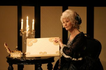 Iäkäs, 1700-luvun asuun pukeutunut nainen istuu pyöreän pöydän ääressä, jossa on kynttelikkö, pitelee koristeellista taulua ja elehtii kuin puhuisi hämärässä, tyylikkäästi sisustetussa huoneessa.