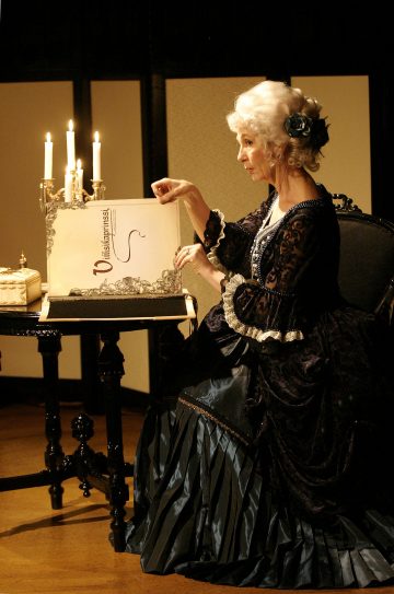 Vanhempi nainen, jolla on koristeellinen, 1700-luvun tyyliin sopiva mekko, istuu pöydän ääressä, jossa on kynttilä, ja osoittaa avoinna olevaa asiakirjaa tai kirjaa, jossa on tyylikäs sisustus ja näkyvissä kakkuja.