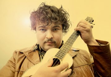 Mies, jolla on kiharat hiukset ja viikset, pukeutuu ruskeaan nahkatakkiin ja pitää kädessään pientä kitaraa tai ukulelea, ja katsoo suoraan kameraan lämpimän keltaista taustaa vasten.