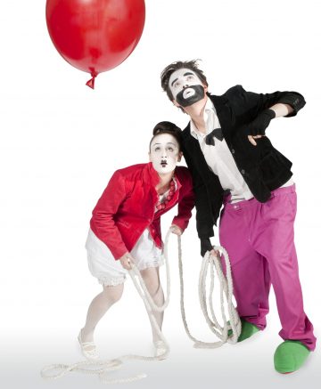 Kaksi valkoiset kasvot meikattua ja ilmeikkäässä asennossa olevaa miimikkoa pitelee köysiä ja katselee yläpuolellaan olevaa punaista ilmapalloa. Toisella on punainen takki ja valkoinen mekko, toisella vaaleanpunaiset housut, musta takki ja vihreät kengät.