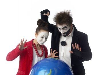 Kaksi valkoisella kasvomaalilla ja mustilla puvuilla varustettua miimikkoa seisoo maapallon takana. Toinen miimikko nostaa nyrkkiä pallon yläpuolelle, kun taas toinen nostaa kätensä ylös yllättyneenä. Molemmilla on teatterimaiset ilmeet valkoista taustaa vasten.