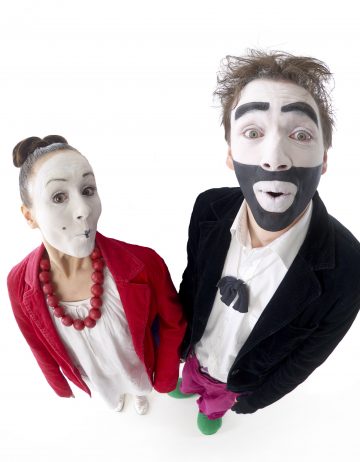 Kaksi miimikkoa seisoo vierekkäin ja katsoo ylöspäin yllättynyt ilme kasvoillaan. Naisella on punainen takki, valkoinen mekko ja punainen kaulakoru. Miehellä on musta takki, rusetti ja rohkea mustavalkoinen kasvomaali.