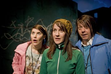 Kolme värikkäisiin takkeihin pukeutunutta ihmistä seisoo lähekkäin tuijottaen eteenpäin yllättynein tai uteliain ilmein. Heidän takanaan näkyy graffitilla päällystetty seinä hämärässä valaistuksessa.