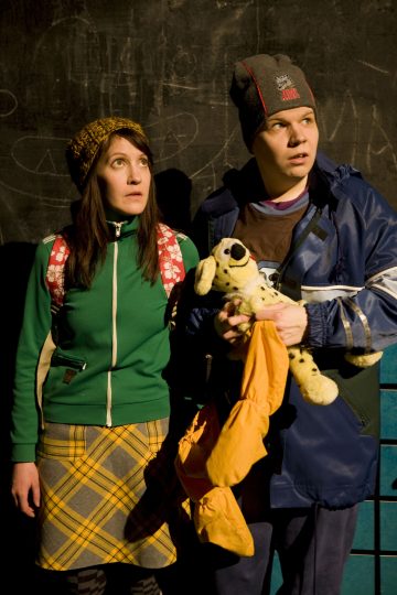 Kaksi ihmistä seisoo liitutaulun edessä ja katsoo ylöspäin yllättyneinä. Toisella on vihreä takki, keltainen ruudullinen hame ja neulomyssy, toisella sininen takki ja harmaa hattu, ja hänellä on pehmolelu ja oranssi laukku.