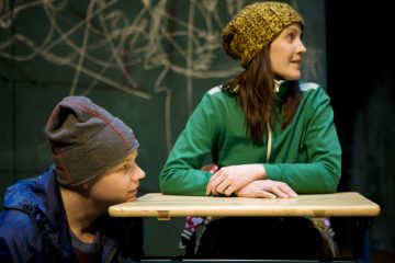 Kaksi pipoon pukeutunutta ihmistä istuu pöydän ääressä. Toinen on kyyristynyt pöydän viereen ja katsoo ylöspäin, kun taas toinen istuu pöydän ääressä ja katsoo sivulle. Taustalla näkyy liitutaulu, johon on raapustuksia.