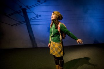 Nuori nainen, jolla on keltainen ruudullinen hame, vihreä takki ja neulomyssy, seisoo dramaattisessa sinisessä valaistuksessa reppu selässään ja käsi ojennettuna puhelinpylväiden muodostaman taustan edessä.