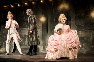 Lavalla esiintyy kolme näyttelijää, joilla on taidokkaat 1700-luvun puvut; kaksi miestä seisoo puhumassa, ja vaaleanpunaiseen pukuun pukeutunut nainen, jolla on kädessään viuhka, istuu tuolilla ja katsoo sivulle. Heidän yläpuolellaan roikkuu valonjohtoja.