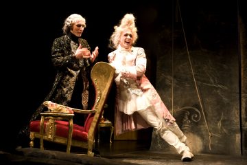 Lavalla esiintyy kaksi näyttelijää koristeellisissa 1700-luvun puvuissa; toisella on musta-kultainen puku ja toisella vaaleanpunainen-valkoinen puku, molemmilla on valkoiset peruukit, ja he seisovat tyylikkään, punapehmusteisen tuolin vieressä dramaattisessa valaistuksessa.