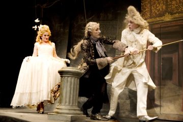 Lavalla esiintyy kolme näyttelijää, jotka ovat pukeutuneet taidokkaisiin 1700-luvun pukuihin. Valkoiseen pukuun pukeutunut nainen hymyilee, kun kaksi miestä, toinen mustiin ja toinen valkoisiin pukeutunut, kamppailevat köyden kanssa kivijalustan lähellä. Lavastus vaikuttaa koristeelliselta ja dramaattiselta.