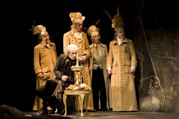 Ryhmä näyttelijöitä, joilla on taidokkaat 1700-luvun puvut ja korkeat peruukit, seisoo pöydän ympärillä, jossa on koristeellisia koristeita, yksi istuu ja muut seisovat, dramaattisessa teatterikohtauksessa, jossa on tumma valaistus.