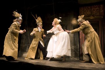 Lavalla esiintyy neljä näyttelijää taidokkaissa puvuissa; kolmella on pitkät ruskeat takit, eläinhatut ja naamiot, kun taas keskellä oleva näyttelijä on pukeutunut valkoiseen mekkoon ja peruukkiin, ja hän vaikuttaa elävältä dramaattisessa kohtauksessa.