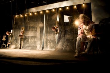 Hämärästi valaistulla näyttämöllä kolme näyttelijää, joilla on taidokkaat aikakauden puvut ja peruukit, kurkistelevat maalatun taustan aukoista, kun taas toinen näyttelijä istuu edessä kyyristyneenä. Näyttämön yläpuolella roikkuu valonjohtoja.