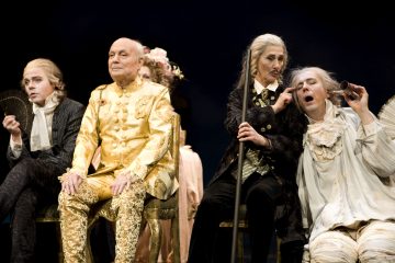 Lavalla istuu neljä näyttelijää taidokkaissa 1700-luvun puvuissa. Yhdellä on kultaa, toinen käyttää viuhkaa, kolmas hoitaa ahdistuneelta vaikuttavan neljännen korvaa. Kohtaus on dramaattinen ja teatraalinen.