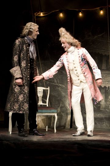 Kaksi näyttelijää näyttämöllä taidokkaissa 1700-luvun puvuissa; toisella on tumma kukkatakki, toisella vaaleanpunainen kirjailtu takki ja valkoiset housut, ja he elehtivät vilkkaasti. Taustalla näkyy valkoinen tuoli ja valonjohtoja.