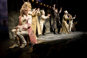 Näyttelijät esiintyvät näyttämöllä taidokkaissa 1700-luvun puvuissa ja peruukissa. Yksi istuu edessä vakavana, kun taas toiset seisovat takana ja elehtivät dramaattisesti näyttämövalojen alla. Kohtaus vaikuttaa teatraaliselta ja dramaattiselta.