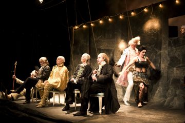 Näyttelijät esiintyvät näyttämöllä taidokkaissa historiallisissa puvuissa, osa istuen ja osa seisten, jousivalojen valossa tummalla, dramaattisella taustalla, jossa on teatterin rekvisiittaa ja lavasteita.