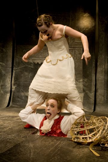 Valkoiseen mekkoon pukeutunut nainen seisoo punaliivinen miehen päällä; hänen päänsä ja kätensä tulevat esiin lattiassa olevasta reiästä. Mies ojentaa leikkisästi kieltään naisen osoittaessa alaspäin, ja lähellä on rikkinäinen kori.