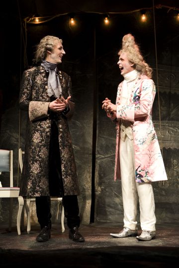 Lavalla seisoo kaksi näyttelijää koristeellisissa 1700-luvun puvuissa hymyillen ja keskustellen. Toisella on tumma kukkatakki ja mustat housut, toisella vaaleanpunainen, kuviollinen takki ja valkoiset housut. Molemmilla on muotoillut peruukit ja dramaattiset ilmeet.
