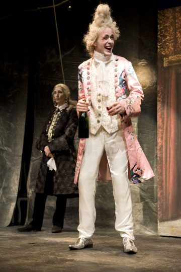 Vaaleanpunaiseen kukkatakkiin, kermanväriseen liiviin ja valkoisiin housuihin pukeutunut henkilö, jolla on puuteroitu peruukki, pitelee pulloa hymyillen lavalla; toinen tummiin aikalaispukuihin pukeutunut henkilö seisoo taustalla.