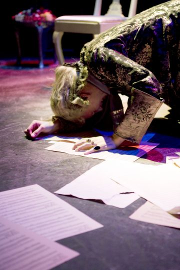 Koristeellisiin vaatteisiin pukeutunut henkilö polvistuu näyttämöllä ja koskettaa otsallaan lattiaa hajallaan olevien paperiarkkien päällä, ja värikäs valaistus korostaa dramaattista kohtausta.