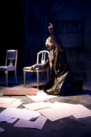 Koristeellisiin vaatteisiin pukeutunut henkilö polvistuu lavalle ja nostaa dramaattisesti yhden käden. Lattiaa peittävät hajallaan olevat paperiarkit, ja kaksi tyhjää tuolia istuu hämärässä, sinisellä valolla valaistussa taustassa.