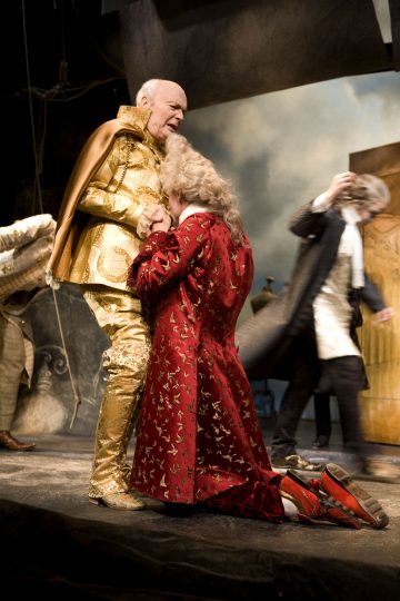 Kultaiseen koristeelliseen pukuun pukeutunut henkilö seisoo, kun toinen, punakuvioinen takki ja valkoinen peruukki yllään, polvistuu ja puristaa kätensä yhteen. Taustalla liikkuu kaksi muuta hahmoa teatterilavalla.