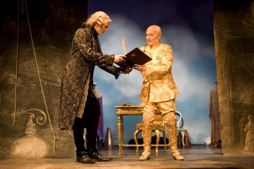 Lavalla esiintyy kaksi näyttelijää koristeellisissa puvuissa; toinen tummassa, kimaltelevassa takissa pitää kirjaa auki toiselle, jolla on hienostunut kultainen puku, ja toinen seisoo pienen pöydän ääressä, jonka taustalla on dramaattinen valaistus ja teatterin rekvisiitta.