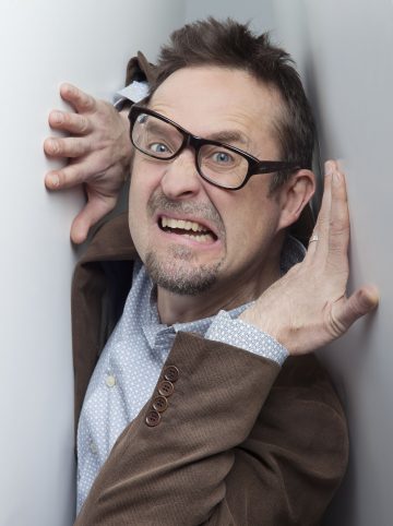 Mies, jolla on silmälasit ja ruskea takki, näyttää säikähtäneeltä ja epämukavalta kahden valkoisen seinän välissä, kädet levällään ja irvistys kasvoillaan.