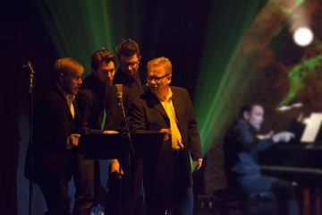 Neljä pukumiestä seisoo lähekkäin ja laulaa lavalla mikrofoniin, kun toinen mies soittaa pianoa taustalla vihreiden lavalamppujen alla.