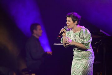 Vihreän ja valkoisen kuviomekkoon pukeutunut nainen laulaa intohimoisesti lavalla mikrofoniin, ja taustalla soittaa violetti valaistus ja epäselvä pianisti.
