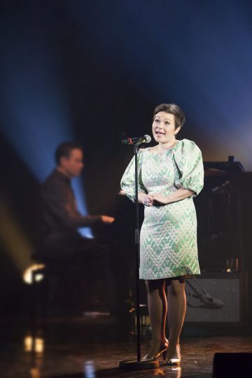 Vihreään kuviomekkoon pukeutunut nainen laulaa lavalla mikrofoniin, ja pianisti soittaa taustalla dramaattisessa valaistuksessa.