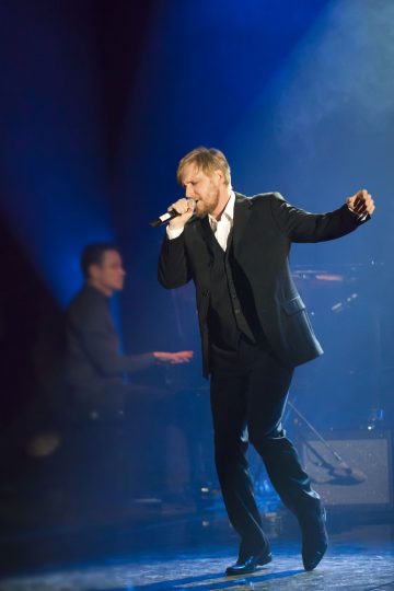 Mustaan pukuun pukeutunut mies laulaa intohimoisesti mikrofoniin lavalla sinisessä valossa, kun toinen muusikko soittaa pianoa taustalla.