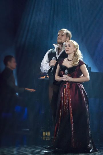 Lavalla on kaksi historiallisiin pukuihin pukeutunutta henkilöä, jotka pitävät mikrofoneja ja laulavat dramaattisessa valaistuksessa. Taustalla näkyy pianisti, joka on osittain epätarkka, mikä lisää esityksen tunnelmaa.
