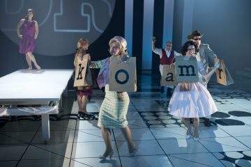 Lavalla on joukko ihmisiä, jotka pitävät kädessään suuria kortteja, joissa on kirjaimet k, o, a, m ja r. Heillä on vintage-tyyliset puvut ja he tanssivat, kun punamekkoon pukeutunut nainen seisoo taustalla korotetulla korokkeella.