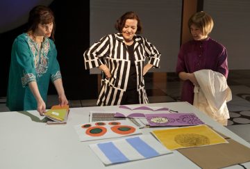 Kolme naista seisoo pöydän ympärillä ja katselee värikkäitä taideteoksia ja papereita, joissa on abstrakteja muotoja ja keltainen ympyrä. Yksi nainen osoittaa, toinen hymyilee, ja kolmas pitelee valkoista liinaa.